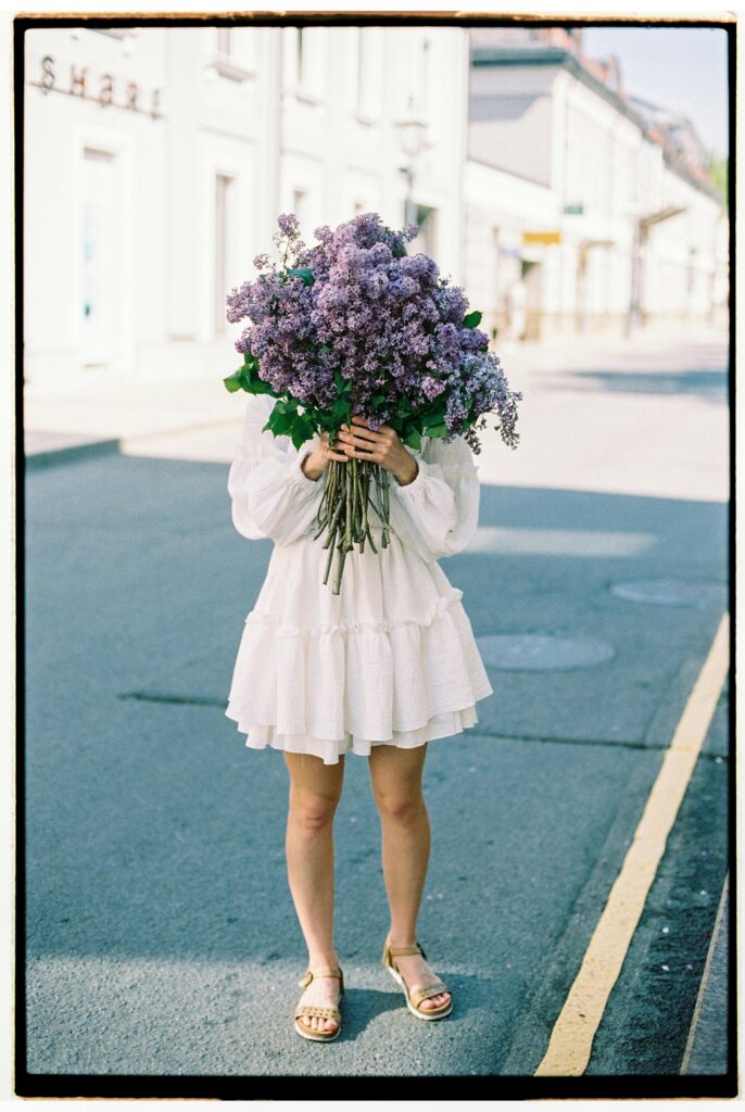 A woman in a white dress holding a bouquet of lilac flowers on an empty street.